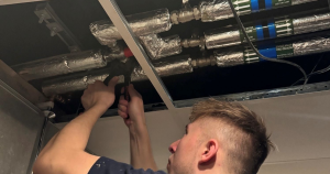 Water hygiene engineer working above a suspended ceiling, inspecting and testing insulated pipework in a building plant system using handheld equipment.