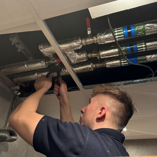 Water hygiene engineer working above a suspended ceiling, inspecting and testing insulated pipework in a building plant system using handheld equipment.